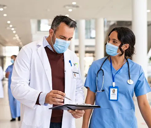 Two medical profesionals wearing white coats and stethoscopes around their necks have a conversation with each other