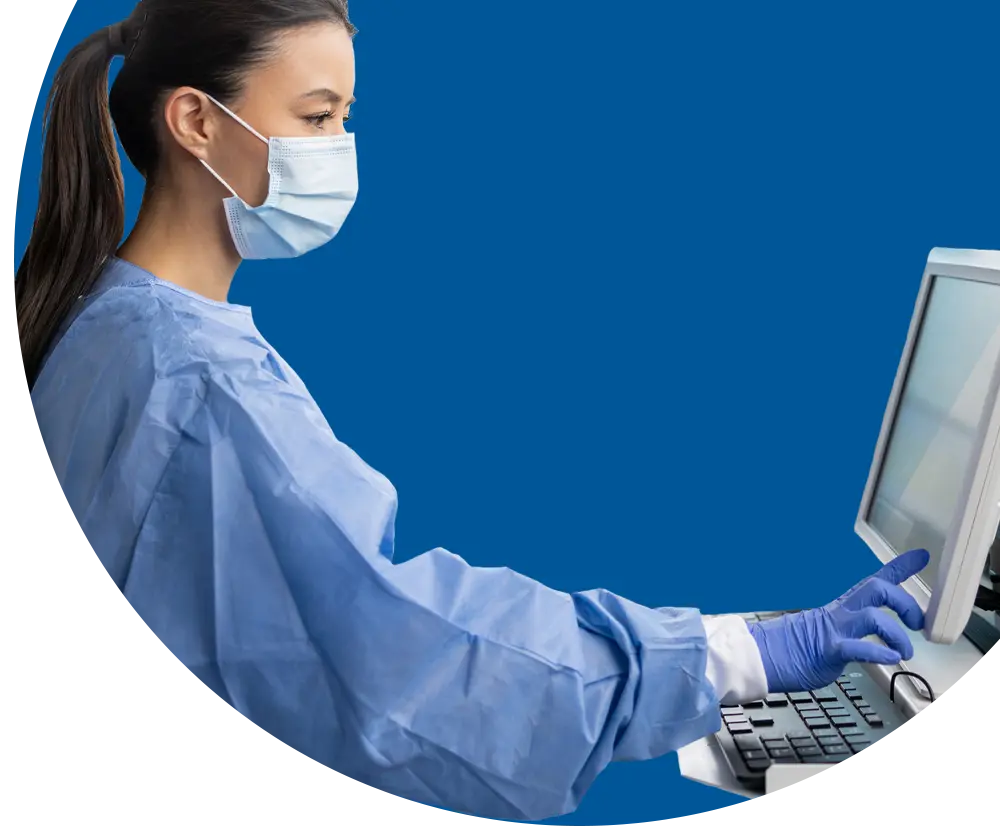 A woman wearing scrubs, a facemask, and latex gloves, uses a touch screen monitor