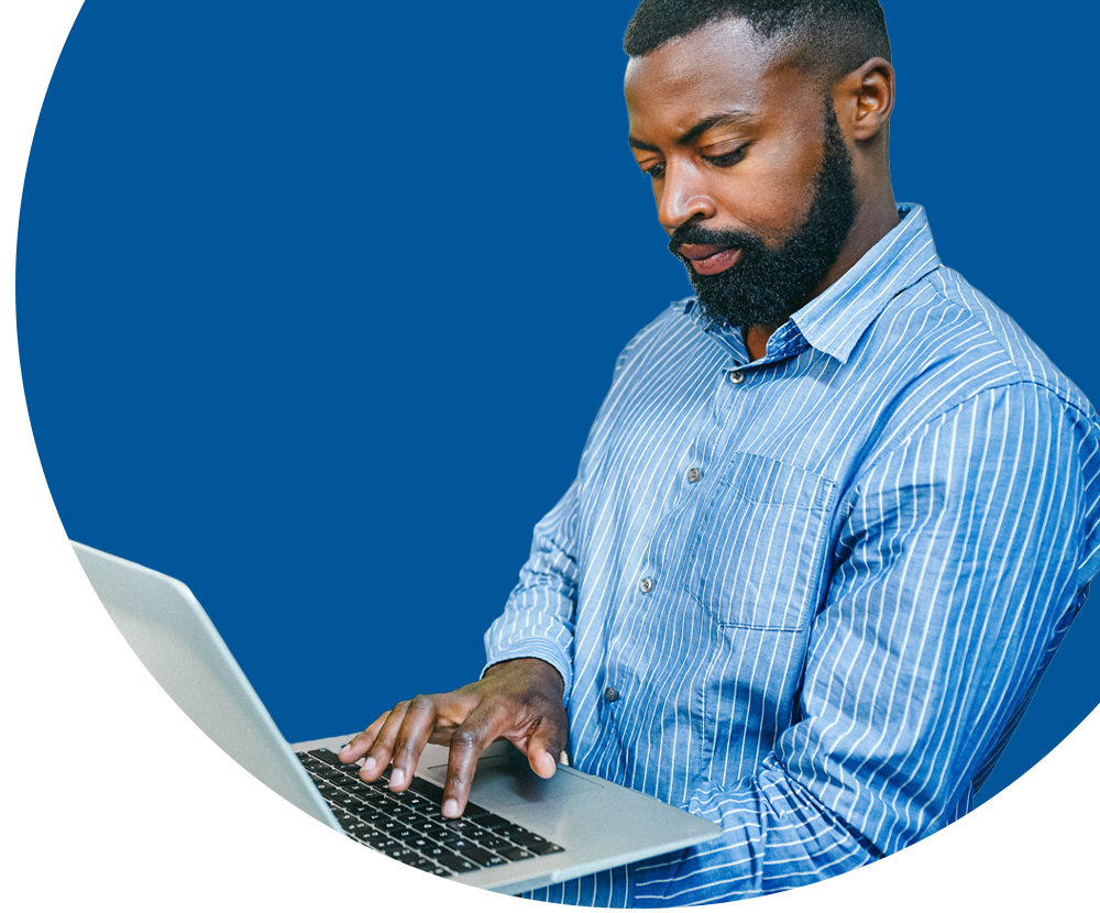 A man with a beard and a blue shirt works on a laptop.