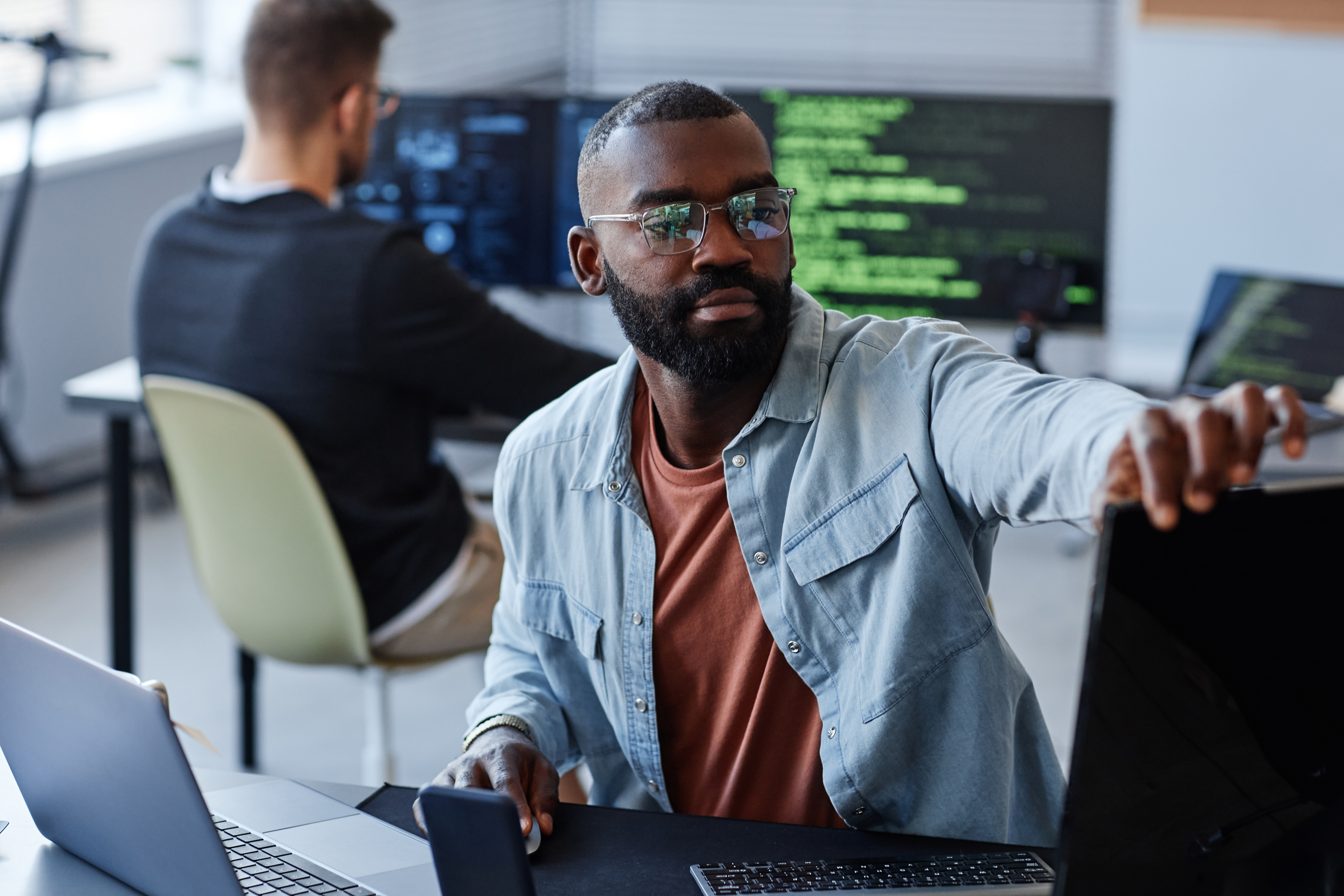 A man with a beard and a blue shirt works on a laptop.